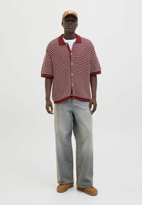 Chemise tricotée à manches courtes dans un motif en vagues rouge et blanc, boutonnée, associée à un jean large gris clair et des chaussures à enfiler beiges.