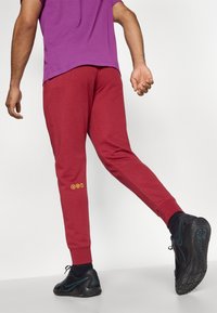 Sarkani sporta bikses ar sašaurinātu piegriezumu, izgatavotas no mīksta auduma; ar dzelteniem logotipa akcentiem labajā potīte un ribotām aprocēm.