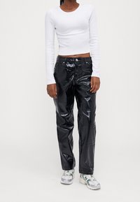 Person som bär en vit långärmad kort topp, glänsande svarta texturerade byxor och vita sneakers med gröna detaljer mot en enfärgad bakgrund.