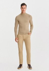 Jasnobrązowy dopasowany sweter z golfem, beżowe spodnie slim-fit i brązowe buty na sznurowadła, na tle jednolitego białego tła.