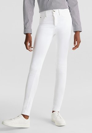 Strakzittende witte jeans gedragen met witte sneakers en een zwart-wit gestreept shirt met lange mouwen op een neutrale achtergrond.