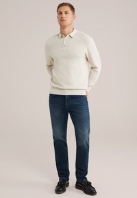 Balts teksturēts džemperis ar apkakli un trīs pogu aizdari, pārī ar tumši ziliem džinsiem un melnām kurpēm, stāvot uz neitrāla fona.