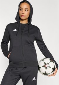 Μαύρο φούτερ με φερμουάρ, κουκούλα με κορδόνια, εμπρόσθιες τσέπες και λευκό λογότυπο Adidas. Συνδυάζεται με μαύρο παντελόνι ίδιου χρώματος και μια μπάλα ποδοσφαίρου με σχέδια.