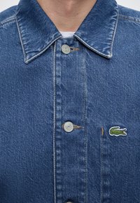 Gros plan d'une veste en jean bleue avec des boutons en métal, un col et un logo crocodile vert sur le côté gauche de la poitrine.
