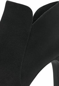 Gros plan sur une chaussure à talon haut en daim noir avec une ouverture pointue et un talon lisse, mettant en valeur la texture et les détails de la couture.