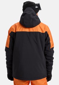 Giacca da sci nera e arancione con cappuccio, maniche lunghe e blocchi di colore a contrasto. Tessuto liscio con materiale impermeabile.