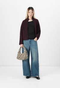 Ung kvinne som har på seg en mørk lilla cardigan, svart topp, vide blå jeans, svarte sko, og holder en beige håndveske med slangeskinnsmønster.