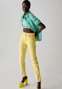Camicia a scacchi verde e bianca sovrapposta a un top corto a righe, abbinata a jeans gialli e tacchi a piattaforma gialli. Sfondo semplice.
