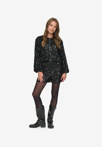 Tenue en sequins noirs comprenant un haut court avec une fermeture éclair et un short assorti. Portée avec des bottes noires jusqu'aux genoux et des collants opaques.
