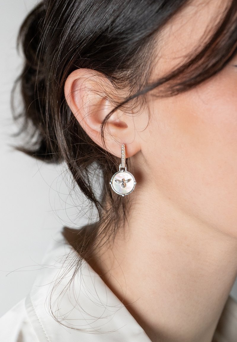 Gros plan sur l’oreille d’une femme portant une boucle d’oreille créole argentée avec un pendentif rond blanc orné d’une petite abeille.