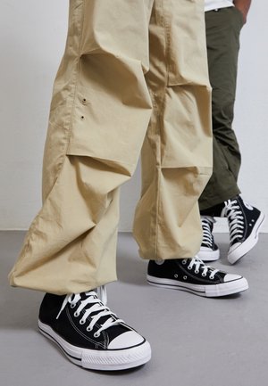 Dos personas que llevan zapatillas negras de caña alta con cordones y suelas blancas, pantalones cargo beige y oliva, de pie sobre un suelo gris.