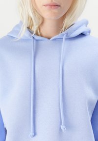 Lyseblå hettegenser med et mykt, teksturert stoff, frontlomme og snorer på hetten, med et avslappet passformdesign.