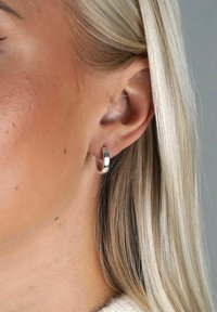 Silver hoopörhänge, slät ytfinish, geometrisk form, rejäl bredd, bärs i örat. Blont hår delvis synligt, neutral bakgrund.