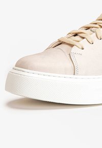 Kerman nilkkuri, jossa on pyöreä kärki, beigellä nauhalla ja tekstuurilla varustettu valkoinen kumipohja. Sivulla näkyy ommeltu yksityiskohtia.