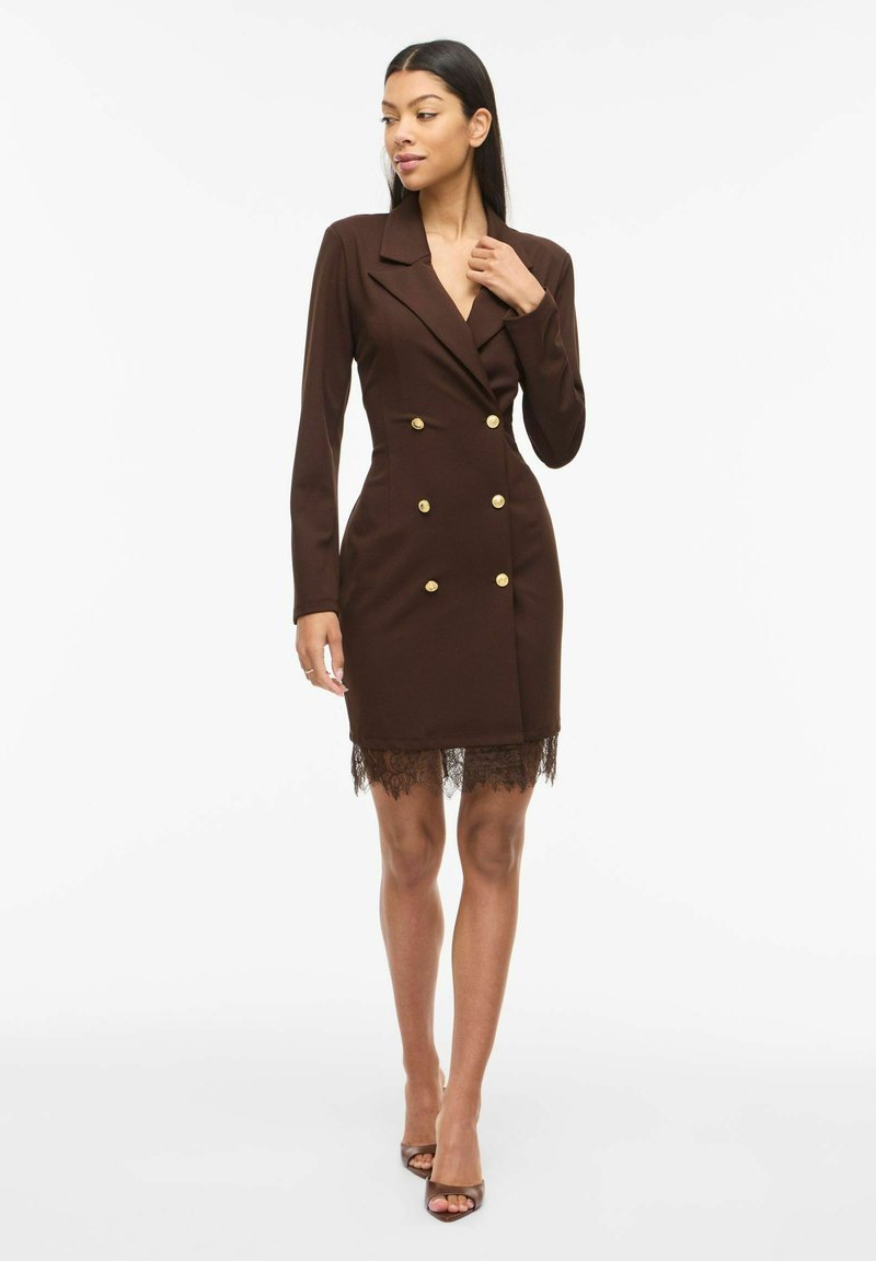 Femme portant une robe blazer marron cintrée à double boutonnage avec des boutons dorés et une bordure en dentelle, associée à des sandales marron à talons.