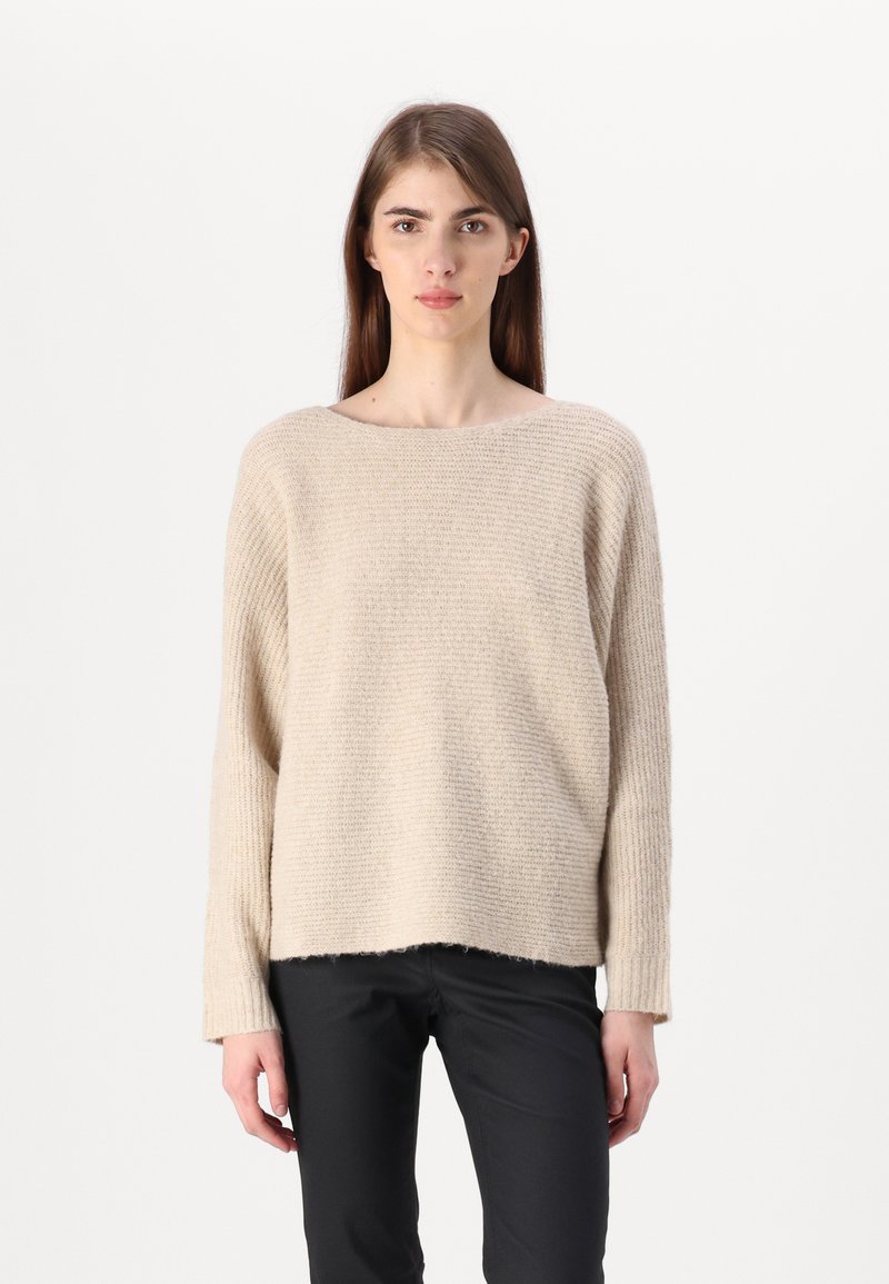 Šviesiai rudos megztinis su plačiu laivagalio apykakle, ilgomis rankovėmis ir šiek tiek trumpesniu šonu. Audinys yra minkštas, su vagotų raštų tekstūra.