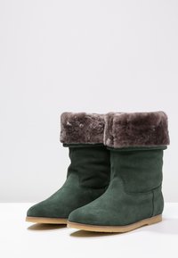 Bottines en daim vert avec un revers en fourrure grise, dotées d'un bout rond et d'une semelle plate couleur tan. Le design est simple et moderne.