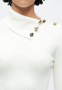 Λευκή ριμπ μπλούζα με ψηλό γιακά, διαγώνιο κλείσιμο με χρυσές διακοσμητικές λεπτομέρειες και υφή καλωδιωτής πλέξης.