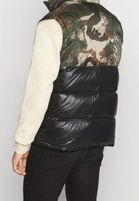 Kamizelka pikowana z górną częścią w camo w odcieniach zieleni i brązu oraz błyszcząca czarna dolna część; zamek błyskawiczny i wysoki kołnierz.