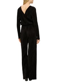 Zwarte jumpsuit met verticale plooien, lange mouwen en een diepe V-rug. De stof lijkt zacht en gestructureerd, met een aansluitende taille en wijde benen.