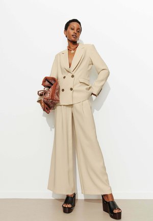 Femme en costume beige à double boutonnage avec pantalon à jambes larges, tenant une écharpe marron à motifs, portant des sandales compensées noires et un collier en forme de soleil.