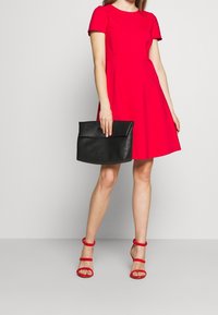 Nainen, jolla on päällään punainen lyhythihainen mekko ja siihen sopivat punaiset korkokengät, pitää suurta mustaa laukkuja vierellään.