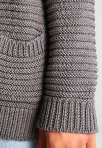 Gray knitted sweater featuring ribbed cuffs and texture varying between smooth and chunky stitches. Includes a front pocket detail.