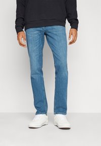 Pantalones de mezclilla azul claro con corte recto, un ligero desgaste y diseño de cinco bolsillos, combinados con zapatillas blancas y una sudadera con capucha negra.