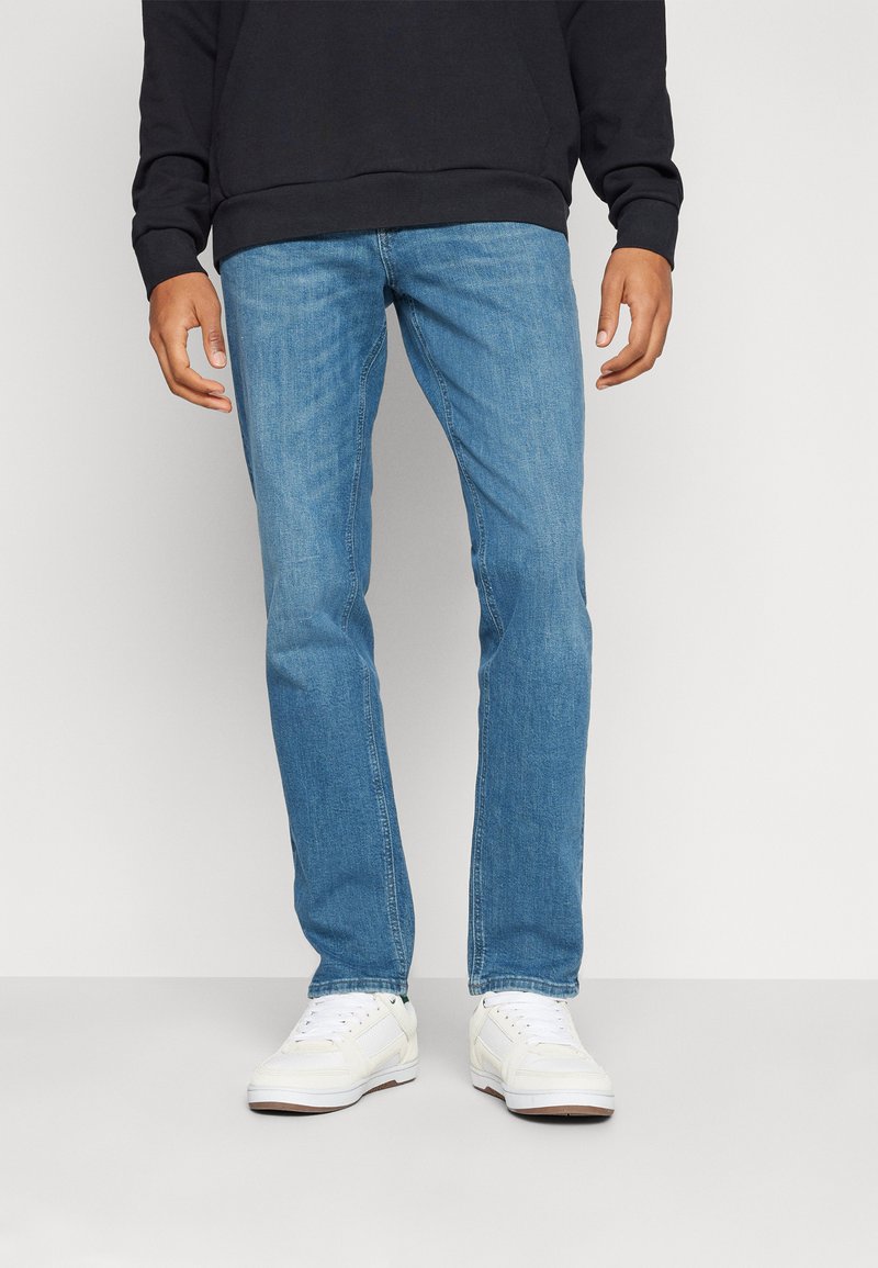 Pantalones de mezclilla azul claro con corte recto, un ligero desgaste y diseño de cinco bolsillos, combinados con zapatillas blancas y una sudadera con capucha negra.