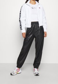 Osoba má na sobě bílou zipovou bundu, zkrácený bílý tričko a černé tepláky, které doplňují barevné tenisky, stojí na neutrálním pozadí.