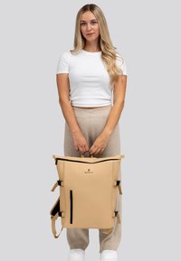 Mujer con camisa blanca y pantalones beige sosteniendo una mochila Pactastic beige por su asa superior frente a un fondo gris claro.
