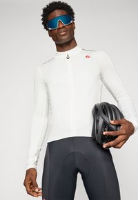 Athlet in weißem Langarm-Radtrikot und schwarzer Radhose hält einen schwarzen Helm und trägt große blaue reflektierende Sportsonnenbrille.