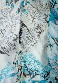 Stoff mit Blumenmuster in Weiß, Schwarz und Türkis. Leichte, durchsichtige Textur mit gefransten Kanten und aufwändigen Mustern.