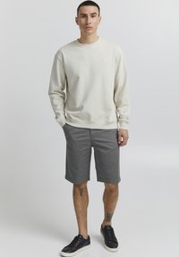 Sudadera cremosa con puños acanalados, combinada con pantalones cortos grises hasta la rodilla. Zapatillas negras completan el conjunto, mostrando un diseño casual.