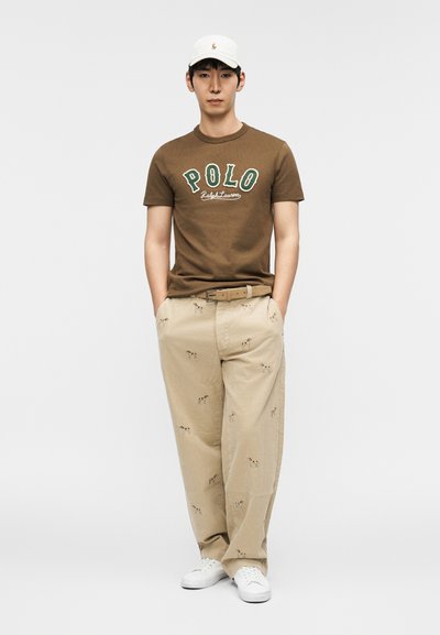 Καφέ μπλουζάκι με λογότυπο "POLO" και κείμενο "Ralph Lauren"; μπεζ παντελόνι με σχέδια σκύλων; λευκά αθλητικά παπούτσια; λευκό καπέλο. Σύνολο casual ένδυσης.