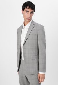 Completo grigio chiaro con motivo a quadri, giacca a un petto con rever a punta, abbinato a una camicia bianca. Il tessuto appare liscio con motivi sottili.