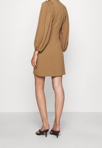 Vestido mini de cuadros marrones con mangas abullonadas, que presenta una tela texturizada. Usado con sandalias negras de tacón alto, mostrado desde atrás.