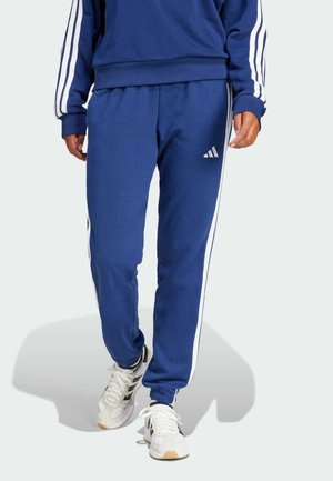 Persoon draagt blauwe Adidas trainingsbroek en sweatshirt met witte strepen, gecombineerd met witte en zwarte sneakers, staand tegen een effen achtergrond.