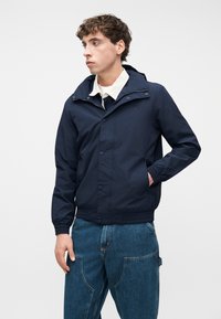 Νέος άνδρας που φοράει μπλε σκούρο μπουφάν με κουκούλα πάνω από λευκό πουκάμισο με γιακά και μπλε τζιν παντελόνι, κοιτάζει προς τα αριστερά με το ένα χέρι στην τσέπη.