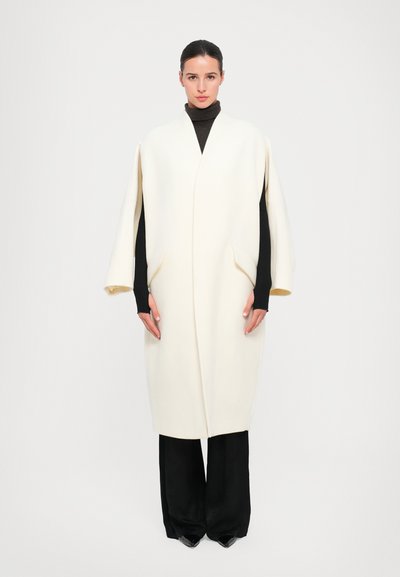 Crème wollen blendjas met een V-hals, ingezakte schouders en zijzakken. Gecombineerd met zwarte broeken en een donkere turtleneck eronder.