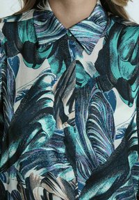 Camisa de cuello con un vibrante patrón abstracto en turquesa, azul marino y púrpura. Tejido suave con cierre de botones y diversos diseños de pinceladas.