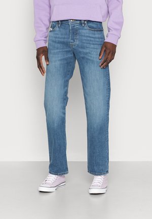 Uomo che indossa jeans azzurri a gamba dritta, felpa con cappuccio lavanda e sneakers basse abbinate di colore lavanda, in piedi contro uno sfondo semplice.