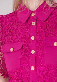 Haut fuchsia avec des détails en dentelle, boutons dorés et deux poches avant. Le design comprend un col et des manches courtes.