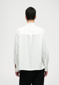 Bílá košile s dlouhým rukávem a volánkovým límcem, horizontálními švy a bočními štěrbinkami na manžetách. Materiál vypadá lehce a hladce.