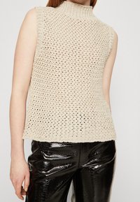 Beige pletený bezrukávový top s vysokým límcem, zdobený texturovaným otevřeným vzorem. Sestaveno s lesklými černými lakovanými kalhotami.