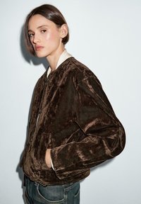 Brun bomberjakke i ruskind med en let skinnende tekstur, der har krave i cremefarve og sidelommer, vist på en neutral baggrund.