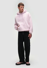 Jasnoróżowa bluza z kapturem i kieszenią kangurką, zestawiona z czarnymi szerokimi spodniami z zamkami oraz wzorzystymi butami.