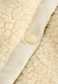 Kermakolorinen kangas, jossa on pehmeä, teksturoitu pinta. Siinä on sileä beige reuna ja pyöreä, kultainen napitusnappi, jossa on kohokuvioitu teksti.