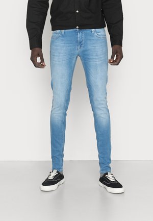 Mand iført lyseblå skinny jeans, sort skjorte med knapper og sorte sneakers med hvide såler, stående mod en ensfarvet hvid baggrund.