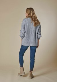 Hellgrauer Strickpullover mit übergroßen Ärmeln, kombiniert mit blauen, schmal zulaufenden Jeans und beigen Ankle-Boots, die einen entspannten Schnitt und eine interessante Textur zeigen.